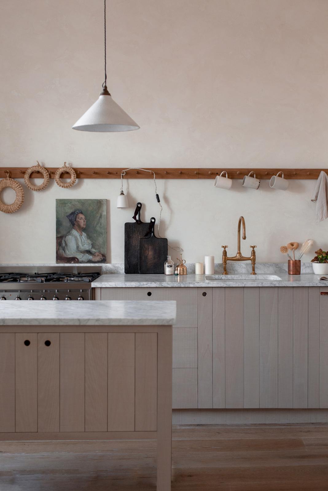 A close-up of a warm and natural Sebastian Cox kitchen features a sink and cooker run with a Carrara marble worktop, a single Belfast sink, an aged brass Ionian tap, a long oak peg rail, and a movable prep table. Beautifully decorated with artisan ceramics, a creamware rise-and-fall light, a plug-in porcelain beaker light, and blackened chopping boards, this kitchen creates a calm and delicate atmosphere that celebrates the use of natural materials.