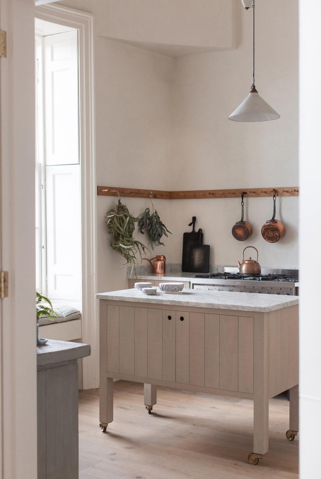 Past a simple doorway is a warm and natural Sebastian Cox kitchen featuring a sink and cooker run with a Carrara marble worktop, a single Belfast sink, an aged brass Ionian tap, a long oak peg rail, and a movable prep table. Beautifully decorated with artisan ceramics, copper pans, drying leaves, and blackened chopping boards, this kitchen creates a calm and delicate atmosphere that celebrates the use of natural materials.