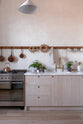 A warm and natural Sebastian Cox kitchen features a sink and cooker run with a Carrara marble worktop, a single Belfast sink, an aged brass Ionian tap, and a long oak peg rail. Beautifully decorated with artisan ceramics, copper pans, delicate potted plants, blackened chopping boards, and a creamware rise-and-fall light, this kitchen creates a calm and delicate atmosphere that celebrates the use of natural materials.