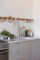 A close-up and oblique view of a warm and natural Sebastian Cox kitchen features a cooker and sink run with a Carrara marble worktop, a single Belfast sink, an aged brass Ionian tap, and a long oak peg rail. Beautifully decorated with artisan ceramics, copper pans, and delicate potted plants, this kitchen creates a calm and delicate atmosphere that celebrates the use of natural materials.