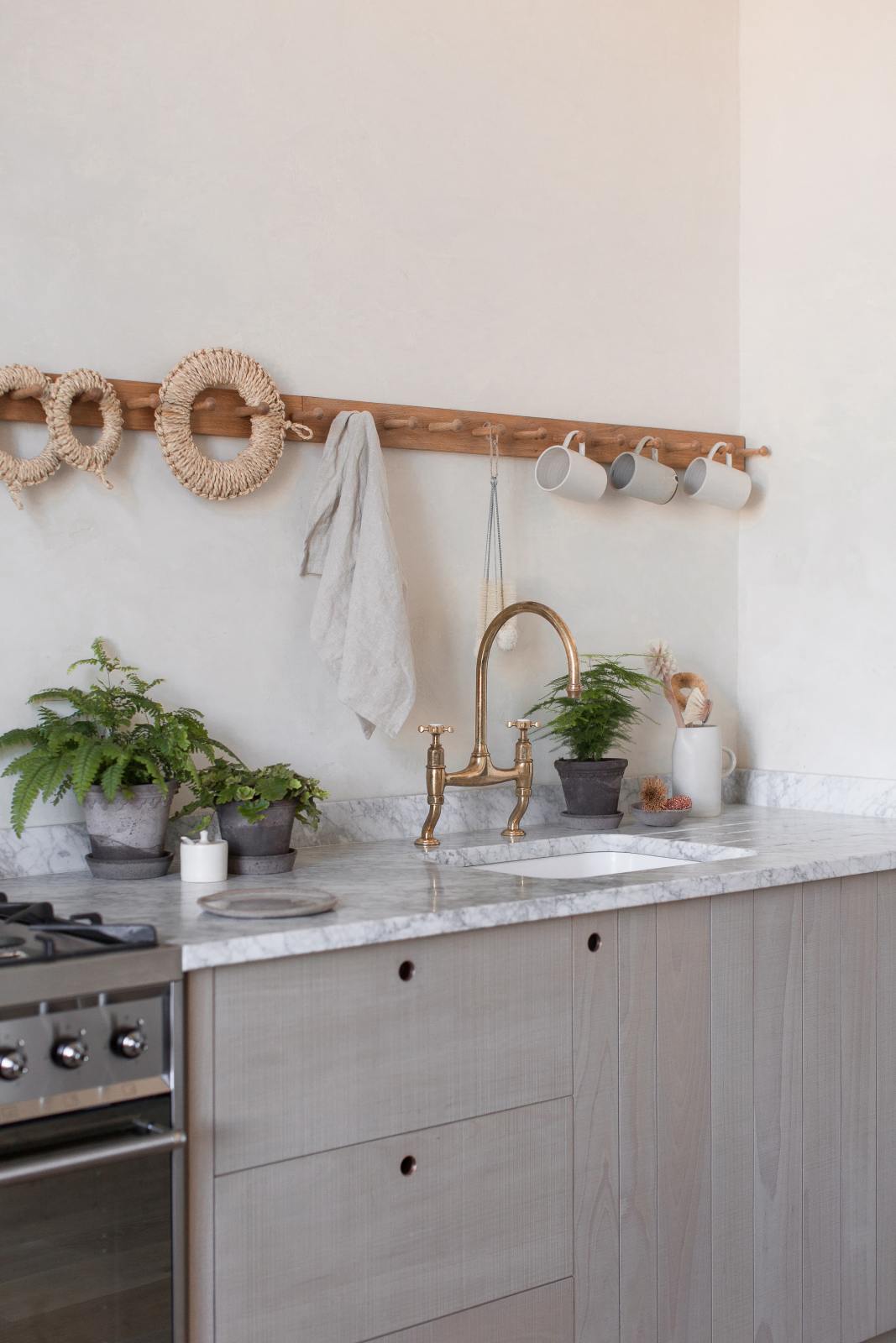 A close-up and oblique view of a warm and natural Sebastian Cox kitchen features a cooker and sink run with a Carrara marble worktop, a single Belfast sink, an aged brass Ionian tap, and a long oak peg rail. Beautifully decorated with artisan ceramics, copper pans, and delicate potted plants, this kitchen creates a calm and delicate atmosphere that celebrates the use of natural materials.