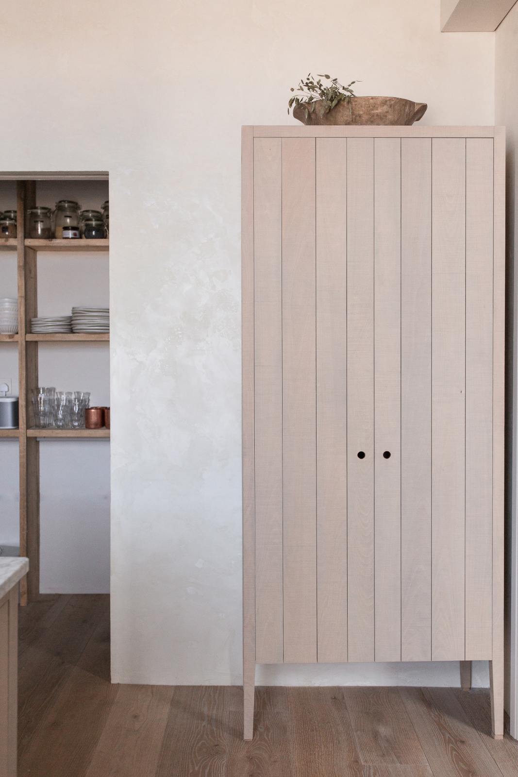 A quiet corner of a warm and natural Sebastian Cox kitchen features a movable prep table with a Carrara marble worktop, a freestanding cupboard, and a walk-in pantry. Beautifully decorated with artisan ceramics, drinking glasses, glass jars with dried ingredients, and a vintage bowl, this kitchen creates a calm and delicate atmosphere that celebrates the use of natural materials.