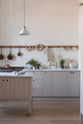A close-up of a warm and natural Sebastian Cox kitchen features a sink and cooker run with a Carrara marble worktop, a single Belfast sink, an aged brass Ionian tap, a long oak peg rail, and a movable prep table. Beautifully decorated with artisan ceramics, a creamware rise-and-fall light, a plug-in porcelain beaker light, and blackened chopping boards, this kitchen creates a calm and delicate atmosphere that celebrates the use of natural materials.