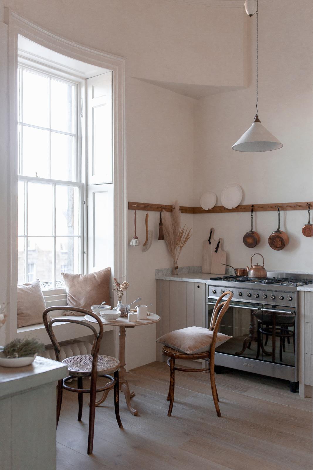 A quiet corner of a warm and natural Sebastian Cox kitchen features a sink and cooker run with a Carrara marble worktop, a single Belfast sink, an aged brass Ionian tap, and a long oak peg rail. Beautifully decorated with vintage furniture, artisan ceramics, a creamware rise-and-fall light, and pale wooden chopping boards, this kitchen creates a calm and delicate atmosphere that celebrates the use of natural materials.