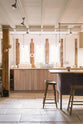 A quiet corner of a soft and spacious Sebastian Cox barn kitchen features a long sink and cooker run with a solid oak worktop, two Belfast sinks, aged brass Mayan taps, a canopy, and a kitchen island with a breakfast nook. Beautifully decorated with lavender colour tints, bare pendant lights, vintage furniture, artisan ceramics, and copper strainers, this kitchen creates an elegant and ethereal space.