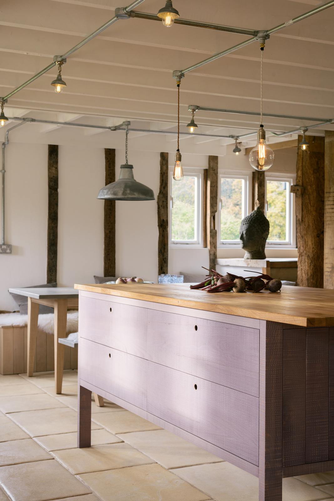 A closer view of a soft and spacious Sebastian Cox barn kitchen island with a breakfast nook. Beautifully decorated with lavender colour tints, bare pendant lights, vintage furniture, artisan ceramics, and organic beets, this kitchen creates an elegant and ethereal space.