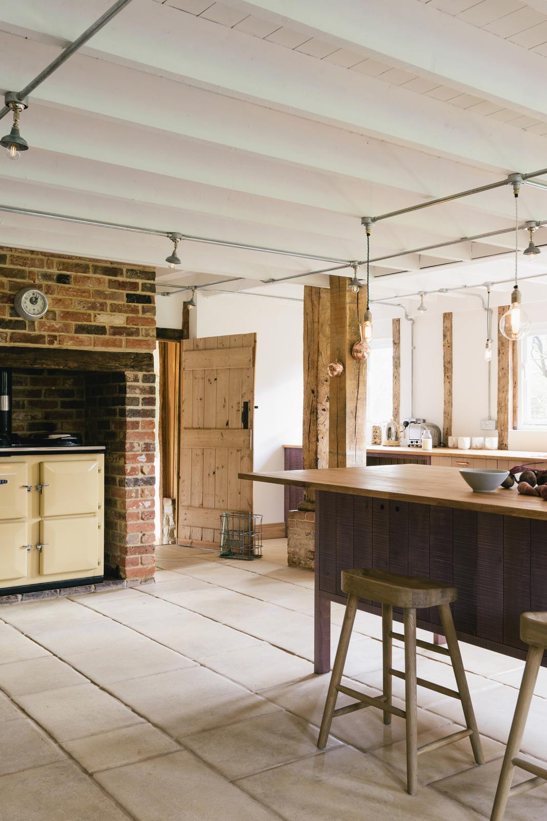 An angled view of a soft and spacious Sebastian Cox barn kitchen features a long sink and cooker run with a solid oak worktop, a kitchen island with a breakfast nook, and a muted yellow gas cooker. Beautifully decorated with lavender colour tints, bare pendant lights, artisan ceramics, wooden stools, and organic beets, this kitchen creates an elegant and ethereal space.