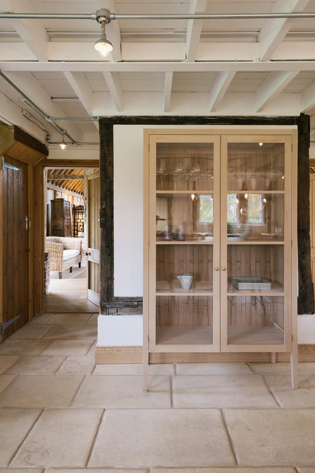On the other side of a soft and spacious Sebastian Cox barn kitchen sits a glazed freestanding cupboard, beautifully decorated with artisan ceramics and drinking glasses. Past a small corridor is a quaint living room featuring rattan armchairs, a vintage wooden partition, and a large oak pantry cupboard.