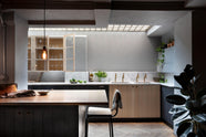 A chic and simple Sebastian Cox basement kitchen features a long sink run with two sets of copper sinks, aged brass Mayan taps, a Carrara marble worktop, glazed wall cupboards, a large kitchen island with a gas cooker, a copper worktop, and a breakfast nook. This kitchen creates a cool and atmospheric space, beautifully decorated with luscious green house plants, vintage items, drinking glasses, a plumber’s stool, and a simple pendant light.