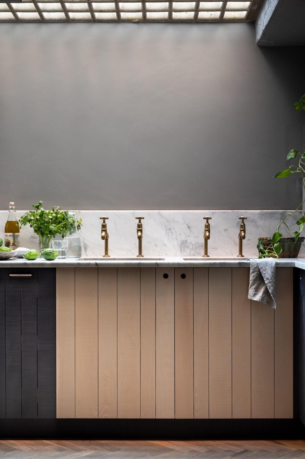 A chic and simple Sebastian Cox basement kitchen features a long sink run with two copper sinks, aged brass Mayan taps, and a Carrara marble worktop and splashback. This kitchen creates a cool and atmospheric space, beautifully decorated with luscious green house plants, vintage items, drinking glasses, a bowl of limes, salt, and parsley.