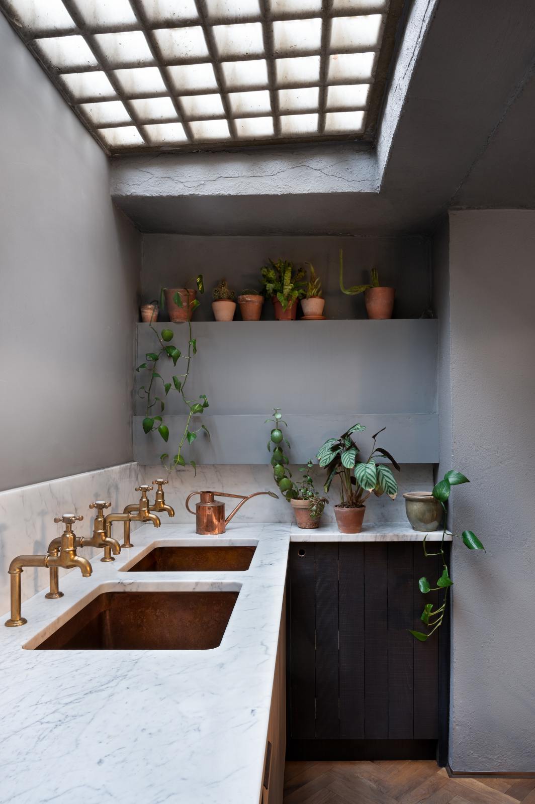 An oblique view of a chic and simple Sebastian Cox basement kitchen features a long sink run with two copper sinks, aged brass Mayan taps, and a Carrara marble worktop and splashback. This kitchen creates a cool and atmospheric space, beautifully decorated with luscious green house plants, a stylish copper watering can, and beautiful ceramic and terracotta pots.