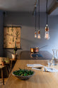 A chic and simple Sebastian Cox basement features a large kitchen island with a gas cooker, a copper and solid oak worktop, and a breakfast nook. This kitchen creates a cool and atmospheric space, beautifully decorated with luscious green cacti, vintage items, a carafe of water, a drinking glass, a bowl of green chillies, and simple pendant lights.