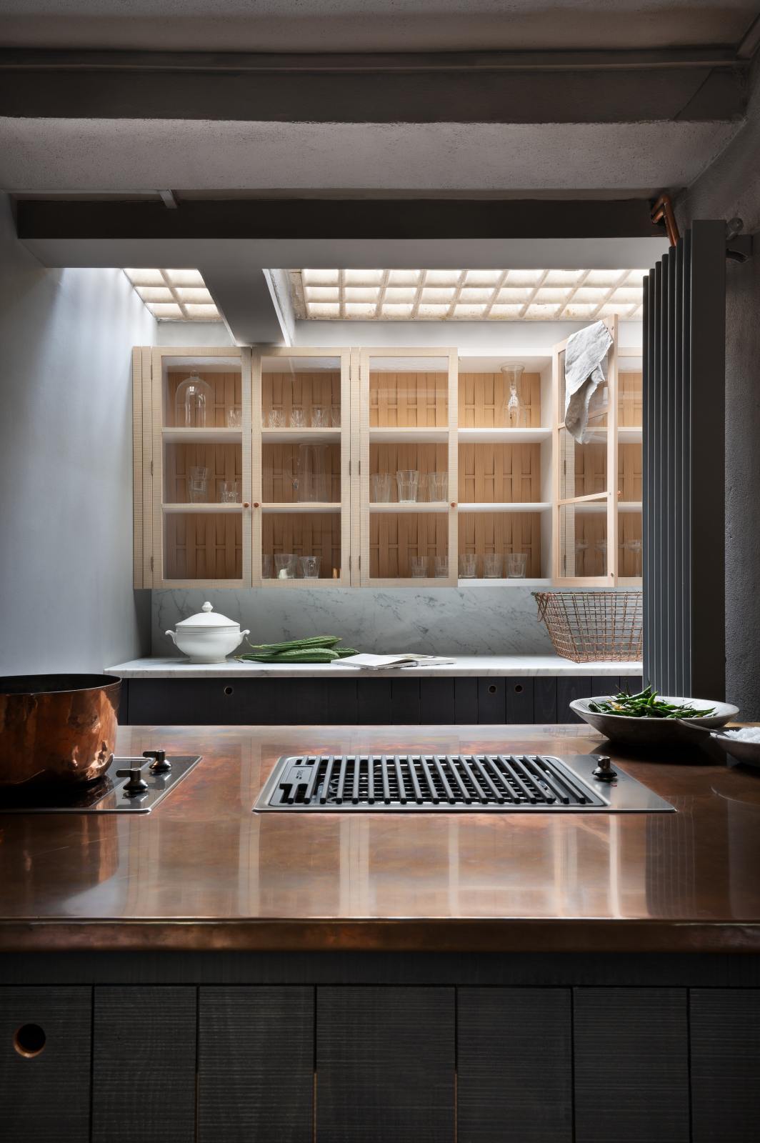 A chic and simple Sebastian Cox basement features a large kitchen island with a gas cooker, a copper worktop, a long sink run with Carrara marble countertop and splashback, and glazed wall cupboards. This kitchen creates a cool and atmospheric space, beautifully decorated with artisan ceramics, a copper pot, a vintage wire basket, drinking glasses, a book, bitter melons, and a bowl of green chillies.