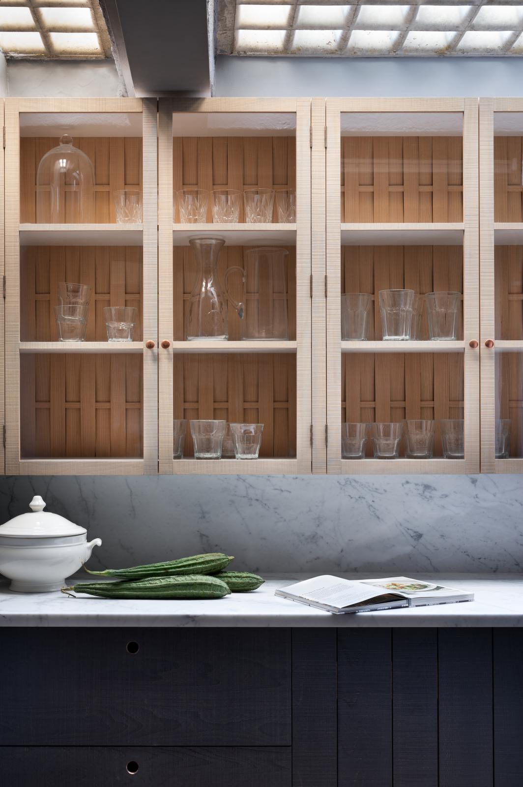 A closer look at a chic and simple Sebastian Cox basement features a long sink run with Carrara marble countertop and splashback, and glazed wall cupboards. This kitchen creates a cool and atmospheric space, beautifully decorated with artisan ceramics, drinking glasses, a book, and bitter melons.