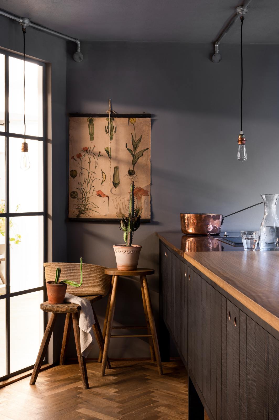 A chic and simple Sebastian Cox basement features a large kitchen island with a gas cooker and a copper and solid oak worktop. This kitchen creates a cool and atmospheric space, beautifully decorated with luscious green cacti, beloved furniture, a Helen stool, a copper pot, a vintage illustration wall-hanging, a carafe of water, a drinking glass, and simple pendant lights.