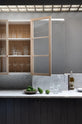 A chic and simple Sebastian Cox basement features a long sink run with two copper sinks, aged brass Mayan taps, and a Carrara marble worktop and splashback. This kitchen creates a cool and atmospheric space, beautifully decorated with olive oil, a carafe of water, drinking glasses, a bowl of limes, a bowl of salt, and books.