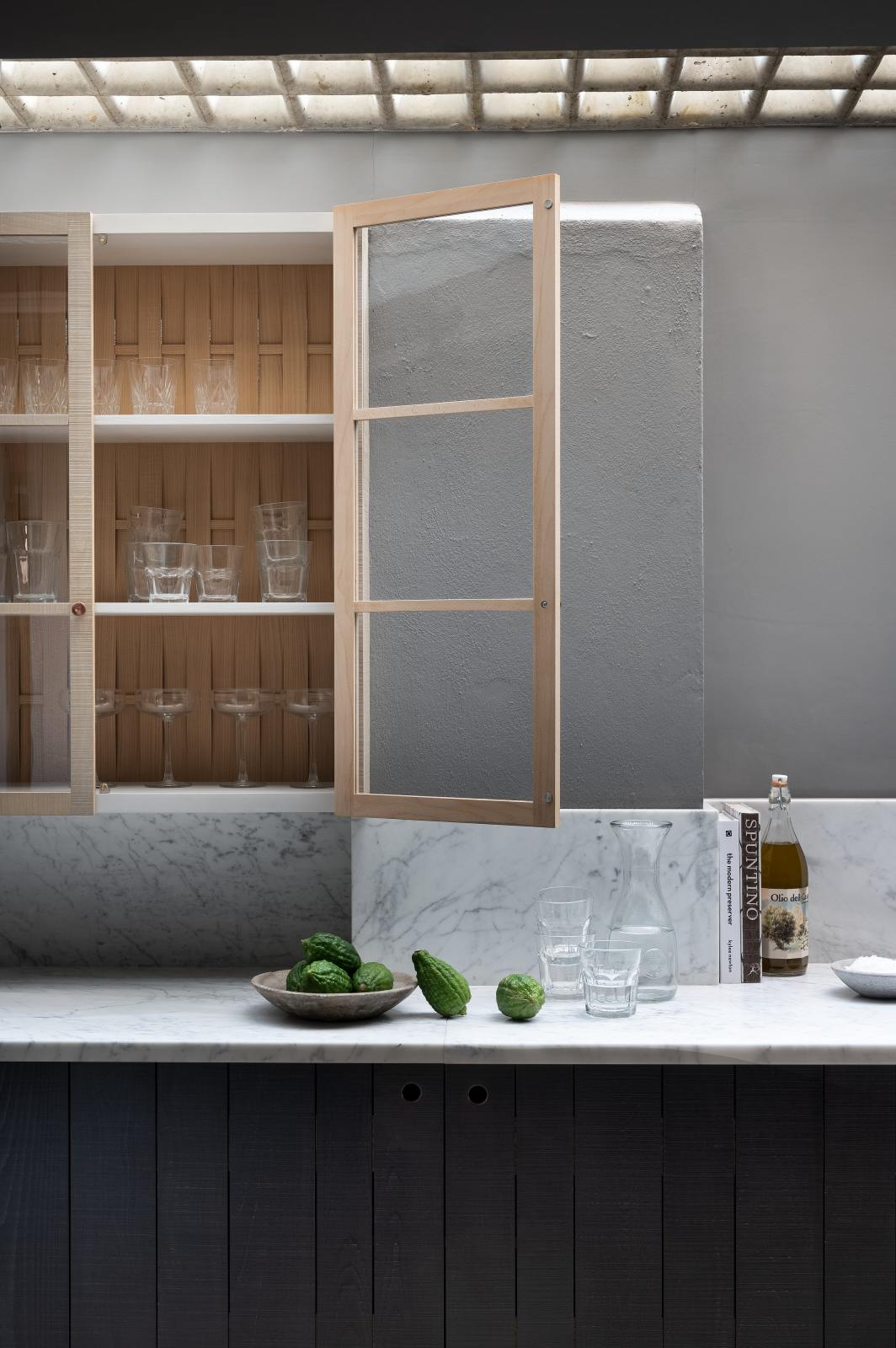 A chic and simple Sebastian Cox basement features a long sink run with two copper sinks, aged brass Mayan taps, and a Carrara marble worktop and splashback. This kitchen creates a cool and atmospheric space, beautifully decorated with olive oil, a carafe of water, drinking glasses, a bowl of limes, a bowl of salt, and books.
