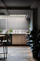 A chic and simple Sebastian Cox basement kitchen features a long sink run with two sets of copper sinks, aged brass Mayan taps, a Carrara marble worktop, glazed wall cupboards, a large kitchen island with a solid oak worktop, and a breakfast nook. This kitchen creates a cool and atmospheric space, beautifully decorated with luscious green house plants, artisan ceramics, a carafe of water, a drinking glass, a plumber’s stool, and a simple pendant light.