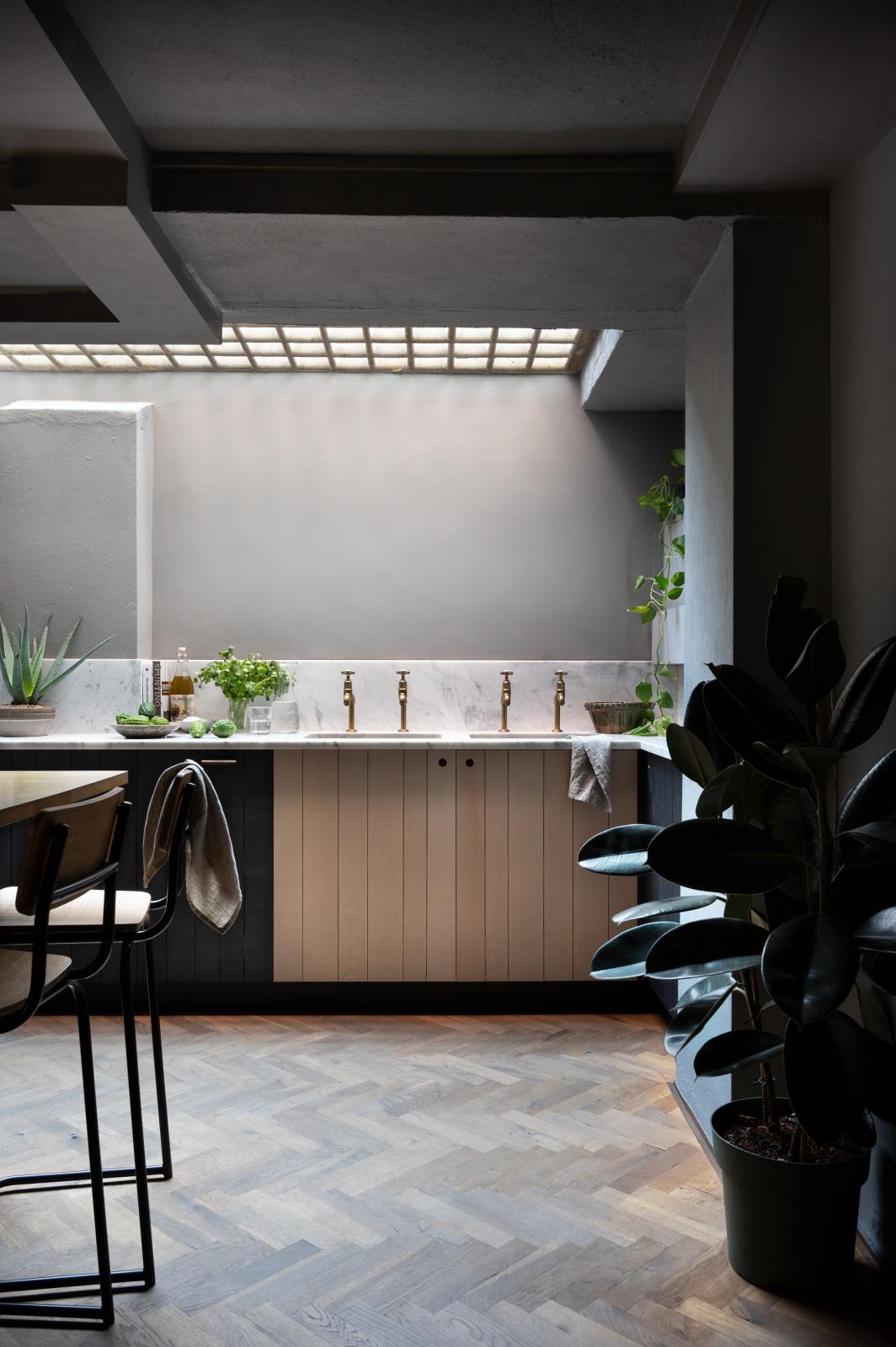 A chic and simple Sebastian Cox basement kitchen features a long sink run with two sets of copper sinks, aged brass Mayan taps, a Carrara marble worktop, glazed wall cupboards, a large kitchen island with a solid oak worktop, and a breakfast nook. This kitchen creates a cool and atmospheric space, beautifully decorated with luscious green house plants, artisan ceramics, a carafe of water, a drinking glass, a plumber’s stool, and a simple pendant light.