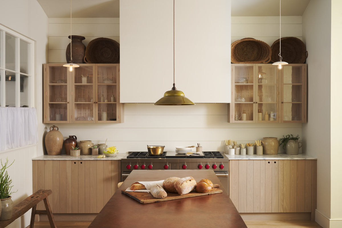 An all-natural and functional Sebastian Cox kitchen features a cooker and sink run with a marble worktop, glazed wall cupboards, and an iconic Haberdasher’s prep table with a copper worktop. Lovingly adorned with antique artisan ceramics, woven baskets, beautiful houseplants, and beloved figurines, it creates a wonderfully simple kitchen with a love of natural sustainability. 