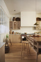 An all-natural and functional Sebastian Cox kitchen features a cooker and sink run with a marble worktop, glazed wall cupboards, and an iconic Haberdasher’s prep table with a copper worktop. Lovingly adorned with antique artisan ceramics, woven baskets, beautiful houseplants, and beloved figurines, it creates a wonderfully simple kitchen with a love of natural sustainability. 