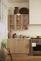 A quiet corner of an all-natural and functional Sebastian Cox kitchen features a cooker and sink run with a marble worktop, glazed wall cupboards, and an iconic Haberdasher’s prep table with a copper worktop. Lovingly adorned with antiques, artisan ceramics, woven baskets, beautiful houseplants, and beloved figurines, it creates a wonderfully simple kitchen with a love of natural sustainability.