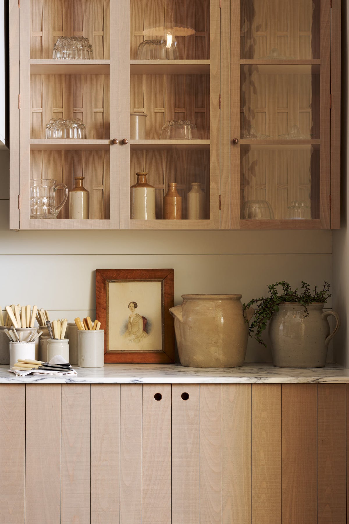 A close-up of an all-natural, functional Sebastian Cox kitchen features a cooker with a marble worktop and glazed wall cupboards. Lovingly adorned with antiques, artisan ceramics, silverware and beautiful houseplants, it creates a wonderfully simple kitchen with a love of natural sustainability.