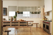 An all-natural and functional Sebastian Cox kitchen features a cooker and sink run with a marble worktop, glazed wall cupboards, and an iconic Haberdasher’s prep table with a copper worktop. Lovingly adorned with antiques, artisan ceramics, cafe curtain rail, and beautiful houseplants, it creates a wonderfully simple kitchen with a love of natural sustainability.