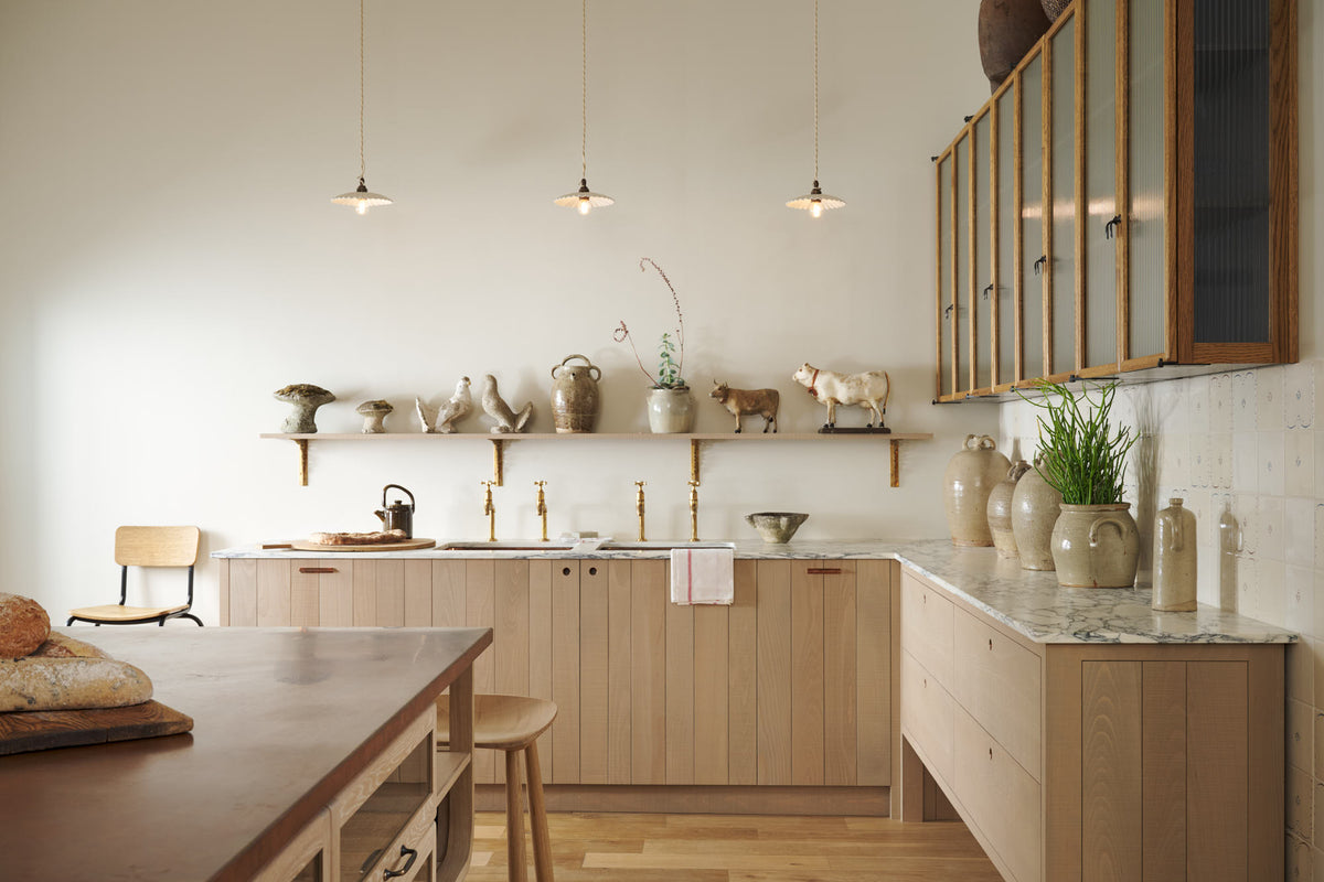 An all-natural and functional Sebastian Cox kitchen features an L-shaped sink run with a marble worktop, glazed wall cupboards, and an iconic Haberdasher’s prep table with a copper worktop. Lovingly adorned with antique artisan pottery, ceramic pendant lights, beautiful houseplants, and beloved figurines, it creates a wonderfully simple kitchen with a love of natural sustainability.