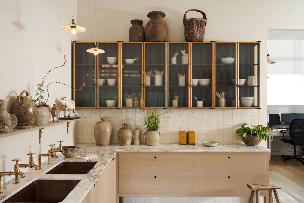 An all-natural and functional Sebastian Cox kitchen features an L-shaped sink run with a marble worktop, glazed wall cupboards, and an iconic Haberdasher’s prep table with a copper worktop. Lovingly adorned with antique artisan pottery, ceramic pendant lights, beautiful houseplants, and beloved figurines, it creates a wonderfully simple kitchen with a love of natural sustainability.