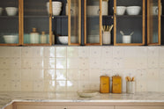 A close-up of an all-natural and functional Sebastian Cox kitchen features an L-shaped sink run with a marble worktop, glazed wall cupboards, and delicate ditsy delft tiles. Lovingly adorned with antique artisan pottery, antiques, and ceramic silverwares, it creates a wonderfully simple kitchen with a love of natural sustainability.