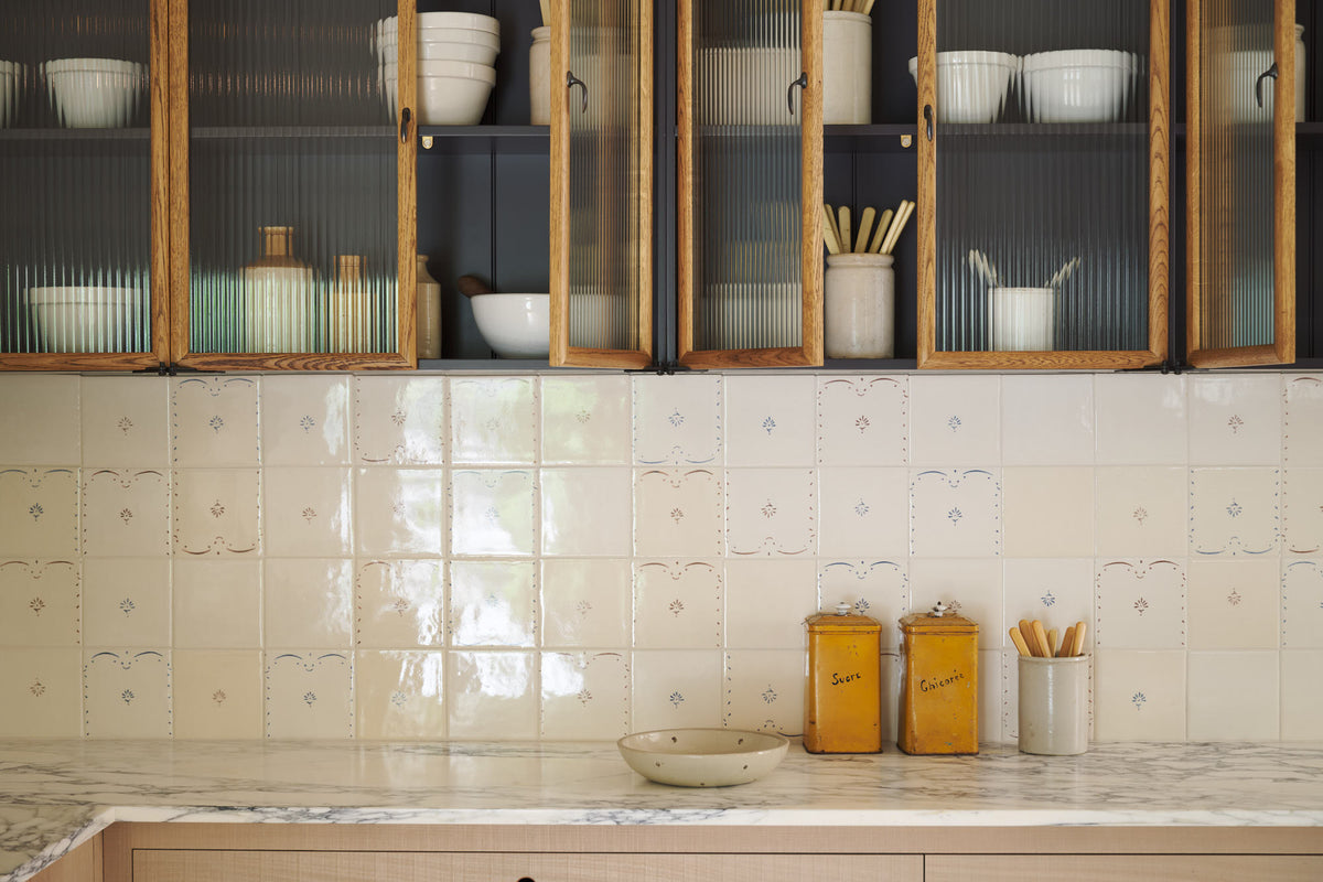 A close-up of an all-natural and functional Sebastian Cox kitchen features an L-shaped sink run with a marble worktop, glazed wall cupboards, and delicate ditsy delft tiles. Lovingly adorned with antique artisan pottery, antiques, and ceramic silverwares, it creates a wonderfully simple kitchen with a love of natural sustainability.
