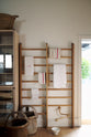 A close-up of an all-natural and functional Sebastian Cox kitchen features a free-standing pantry with glazed doors, an aged brass pot filler tap, and a tall Haberdasher’s hanging rack. Lovingly adorned with antique artisan pottery, woven baskets, and tea towels, it creates a wonderfully simple kitchen with a love of natural sustainability.