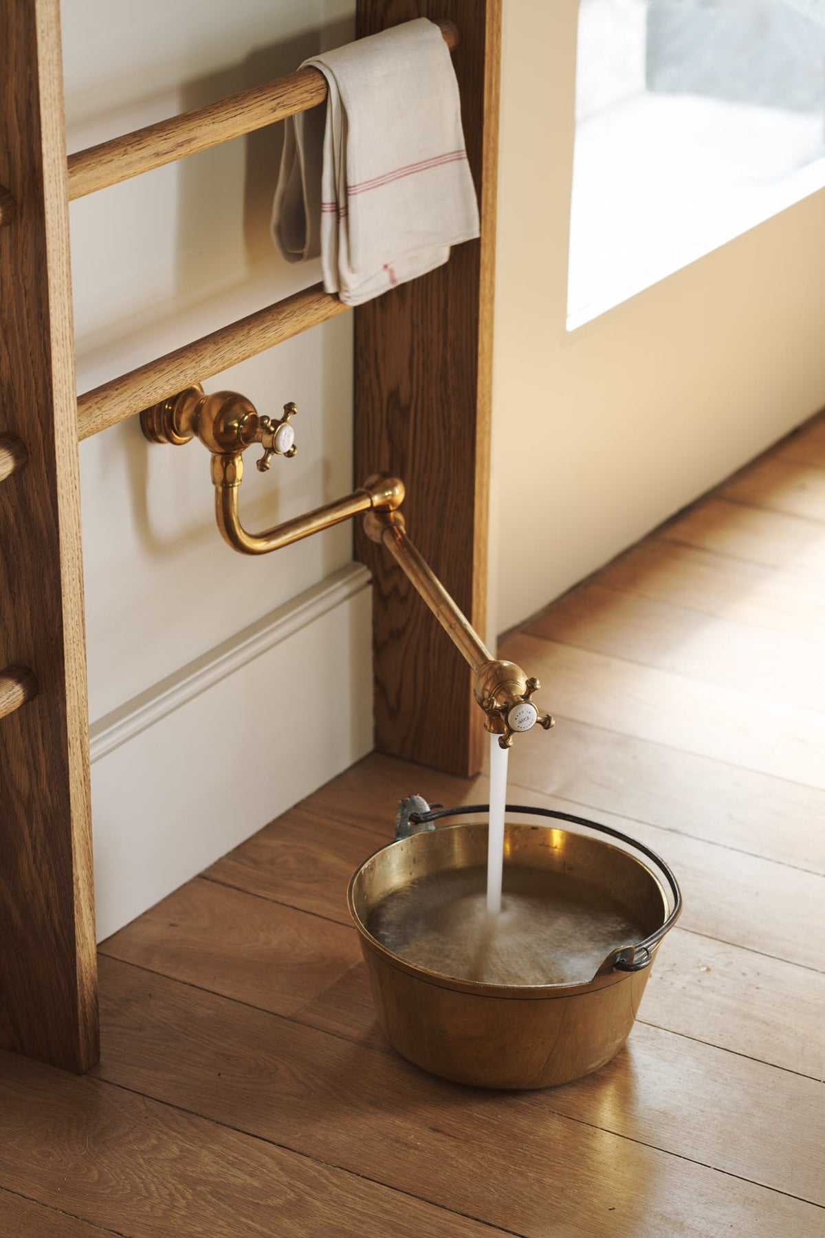 A close-up of an all-natural and functional Sebastian Cox kitchen features an aged brass pot filler tap and a tall Haberdasher’s hanging rack. Lovingly adorned with a tea towel and a brass pan for a makeshift dog bowl.