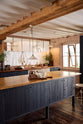 An oblique view of a warm and stylish Sebastian Cox kitchen features a sink run with two single copper sinks, aged brass Mayan taps, glazed partitions, and a copper worktop. Adorned with an aged brass café curtain rail, a milk glass task light, a creamware rise and fall light, artisan ceramics, and vintage wooden items, this kitchen creates a humble space that celebrates a love of simple and natural materials.
