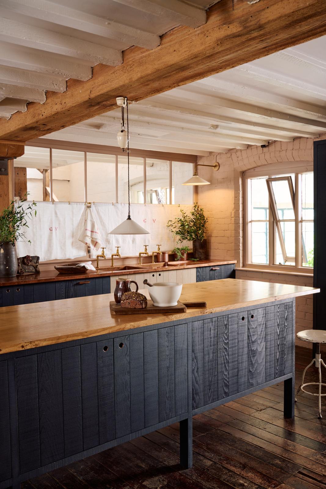 An oblique view of a warm and stylish Sebastian Cox kitchen features a sink run with two single copper sinks, aged brass Mayan taps, glazed partitions, and a copper worktop. Adorned with an aged brass café curtain rail, a milk glass task light, a creamware rise and fall light, artisan ceramics, and vintage wooden items, this kitchen creates a humble space that celebrates a love of simple and natural materials.