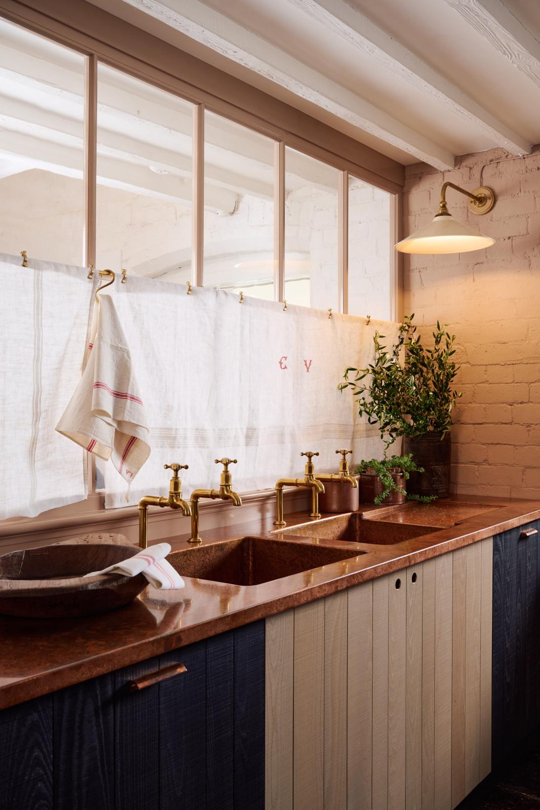 An oblique view of a warm and stylish Sebastian Cox kitchen features a sink run with two single copper sinks, aged brass Mayan taps, glazed partitions, and a copper worktop. Adorned with an aged brass café curtain rail, a milk glass task light, artisan ceramics, and vintage wooden items, this kitchen creates a humble space that celebrates a love of simple and natural materials.