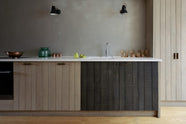 The sleek and simple Sebastian Cox kitchen includes a cooker and sink run topped with a Silestone worktop, a full-height larder, a single Belfast sink, a silver tap, and walls finished with natural limestone. The space is beautifully decorated with a plate of fresh green pears, red-tinted drinking glasses, copper shot glasses, a large blue vase, and a copper bowl, creating a minimal yet earth-conscious and sustainable kitchen environment.
