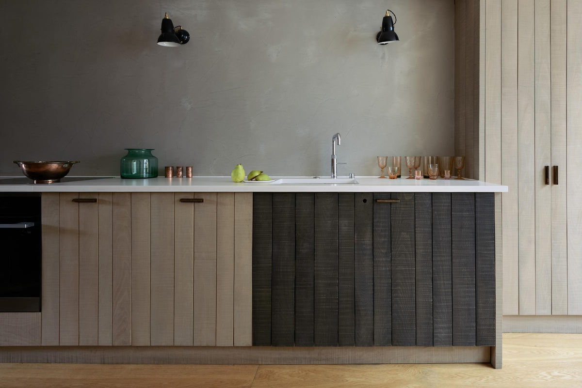 The sleek and simple Sebastian Cox kitchen includes a cooker and sink run topped with a Silestone worktop, a full-height larder, a single Belfast sink, a silver tap, and walls finished with natural limestone. The space is beautifully decorated with a plate of fresh green pears, red-tinted drinking glasses, copper shot glasses, a large blue vase, and a copper bowl, creating a minimal yet earth-conscious and sustainable kitchen environment.