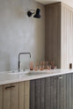 An oblique view of a sleek and simple Sebastian Cox kitchen includes a cooker and sink run topped with a Silestone worktop, a full-height larder, a single Belfast sink, a silver tap, and walls finished with natural limestone. The space is beautifully decorated with a plate of fresh green pears, red-tinted drinking glasses, copper shot glasses, a large blue vase, and a copper bowl, creating a minimal yet earth-conscious and sustainable kitchen environment.