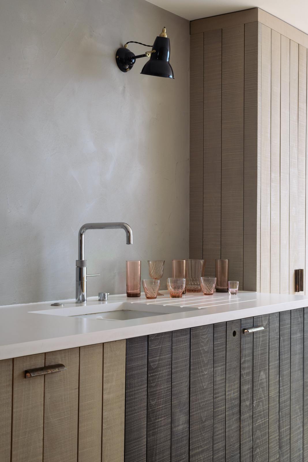 An oblique view of a sleek and simple Sebastian Cox kitchen includes a cooker and sink run topped with a Silestone worktop, a full-height larder, a single Belfast sink, a silver tap, and walls finished with natural limestone. The space is beautifully decorated with a plate of fresh green pears, red-tinted drinking glasses, copper shot glasses, a large blue vase, and a copper bowl, creating a minimal yet earth-conscious and sustainable kitchen environment.