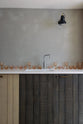 A close-up of a sleek and simple Sebastian Cox kitchen includes a cooker and sink run topped with a Silestone worktop, a full-height larder, a single Belfast sink, a silver tap, and walls finished with natural limestone. The space is beautifully decorated with red-tinted drinking glasses and a black industrial wall lamp, creating a minimal yet earth-conscious and sustainable kitchen environment.