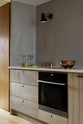 An oblique view of a sleek and simple Sebastian Cox kitchen includes a cooker and sink run topped with a Silestone worktop, and walls finished with natural limestone. The space is beautifully decorated with a plate of fresh green pears, copper shot glasses, a copper bowl, and a black industrial wall lamp, creating a minimal yet earth-conscious and sustainable kitchen environment.