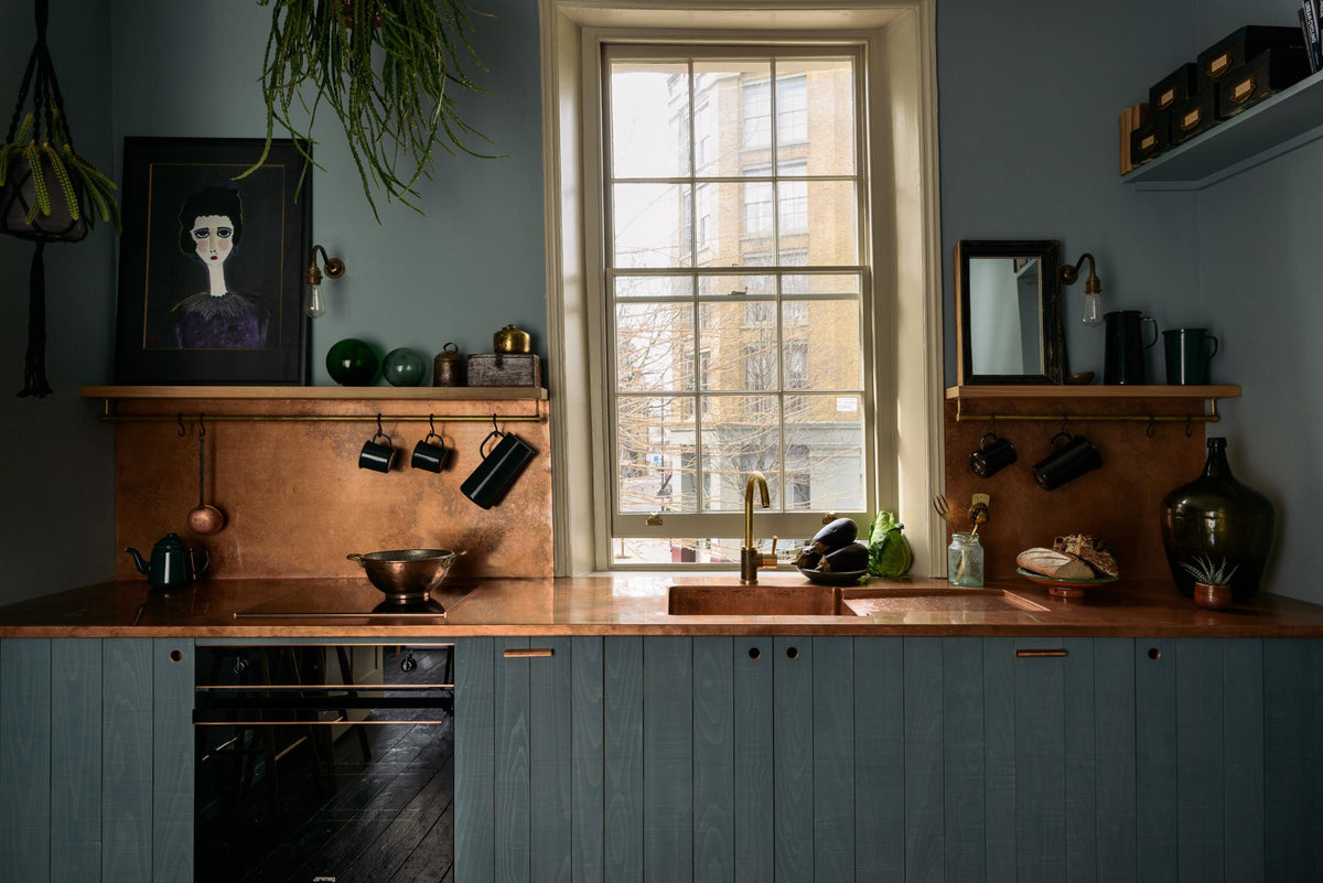 A sweet and simple Clerkenwell blue Sebastian Cox kitchen features a short cooker and sink run with a copper worktop and splashback, a single copper sink, an electric cooker, and shelves with brass hanging rails. Adorned with artisan ceramics, vintage bric-a-brac, aubergines, a sweetheart lettuce, loaves of bread, hanging plants, a framed quirky illustration, and bare wall lights, this kitchen creates a glamorous and practical space.
