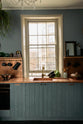 A close-up of a sweet and simple Clerkenwell blue Sebastian Cox kitchen features a short cooker and sink run with a copper worktop and splashback, a single copper sink, an electric stove, and shelves with brass hanging rails. Adorned with artisan ceramics, vintage bric-a-brac, aubergines, a sweetheart lettuce, loaves of bread, hanging plants, and a bare wall light, this kitchen creates a glamorous and practical space.