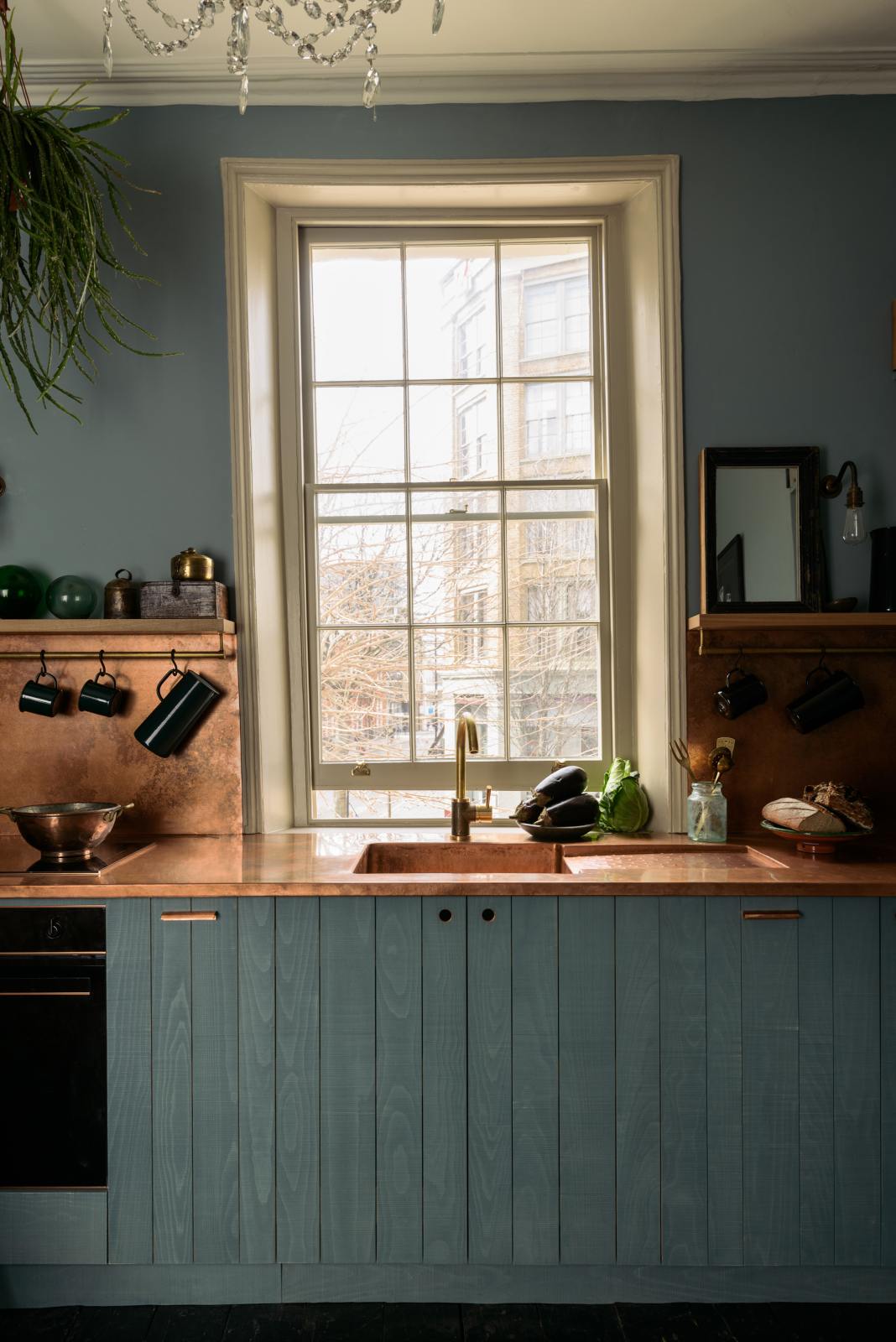A close-up of a sweet and simple Clerkenwell blue Sebastian Cox kitchen features a short cooker and sink run with a copper worktop and splashback, a single copper sink, an electric stove, and shelves with brass hanging rails. Adorned with artisan ceramics, vintage bric-a-brac, aubergines, a sweetheart lettuce, loaves of bread, hanging plants, and a bare wall light, this kitchen creates a glamorous and practical space.