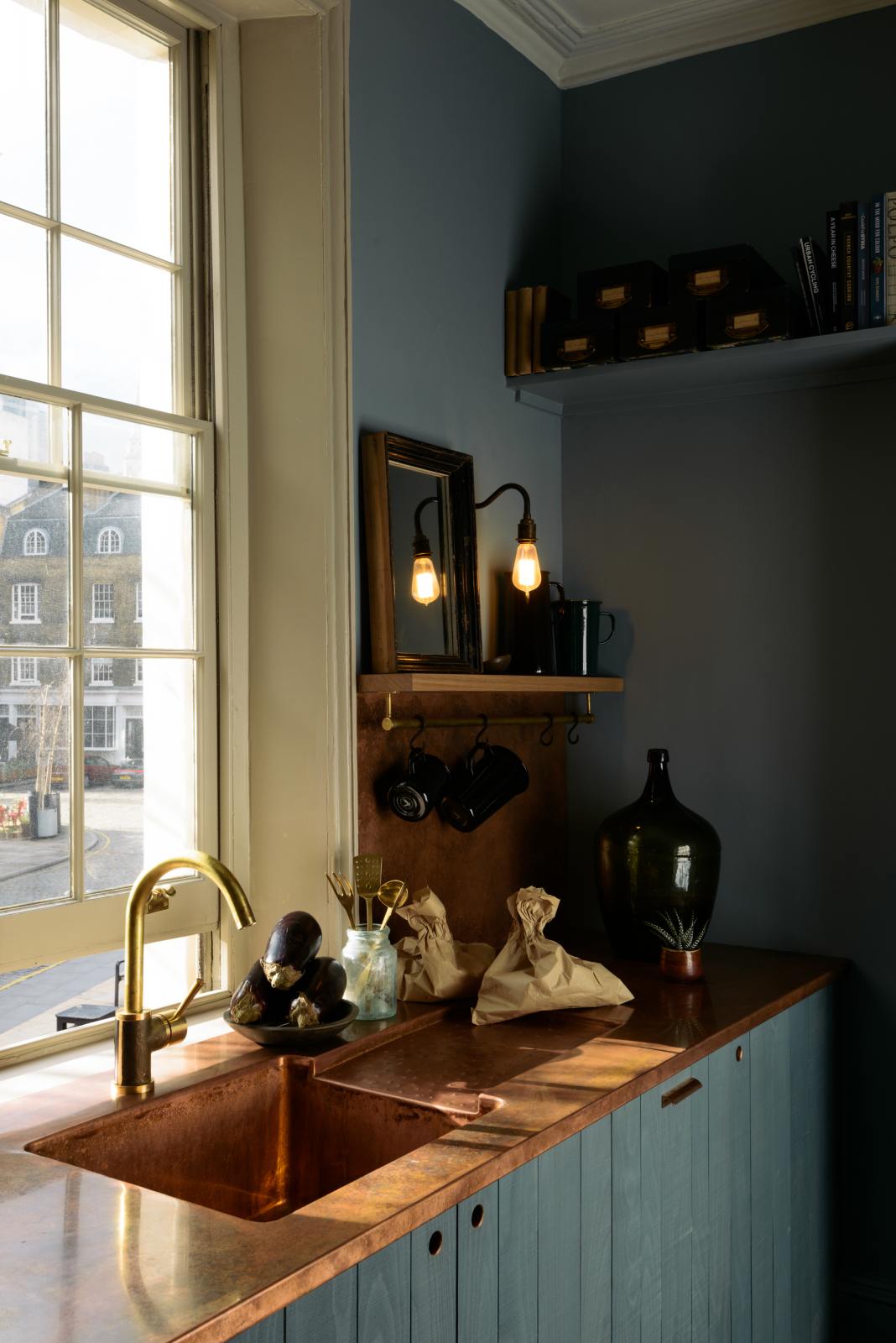 An oblique view of a sweet and simple Clerkenwell blue Sebastian Cox kitchen features a short cooker and sink run with a copper worktop and splashback, a single copper sink, and shelves with brass hanging rails. Adorned with artisan ceramics, vintage bric-a-brac, aubergines, hanging plants, and a bare wall light, this kitchen creates a glamorous and practical space.