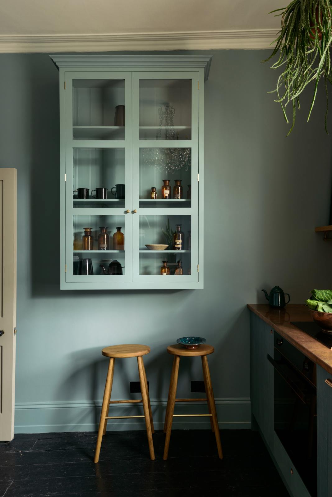 A sweet and simple Clerkenwell blue Sebastian Cox kitchen features a short cooker and sink run with a copper worktop and splashback, and a glazed wall cupboard. Adorned with artisan ceramics, vintage bric-a-brac, hanging plants, a framed quirky illustration, and Helen stools, this kitchen creates a glamorous and practical space.