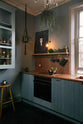 An angled view of a sweet and simple Clerkenwell blue Sebastian Cox kitchen features a short cooker and sink run with a copper worktop and splashback, a single copper sink, an electric stove, shelves with brass hanging rails, and a glazed wall cupboard. Adorned with artisan ceramics, vintage bric-a-brac, hanging plants, a sweetheart cabbage, a framed quirky illustration, and a bare wall light, this kitchen creates a glamorous and practical space.