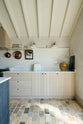 A quiet corner of a beautifully renovated farm features a Sebastian Cox kitchen with a long cooker run, a large kitchen island with a double fluted Belfast sink, an aged brass Ionian tap, and a Carrara marble and solid oak worktop. Adorned with Heirloom wall lights, vintage items, artisan ceramics, and an oil painting, this kitchen creates a clean and contemporary style that harmonises with the natural materials used throughout the space.